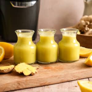 Healthy homemade ginger shots made with fresh ginger, pineapple, and lemon juice in three small glass bottles. Surrounded by fresh ginger root, lemons, and a juicer, these immune-boosting wellness shots are perfect for meal prep and daily nutrition.