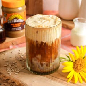 Creamy iced SunButter latte topped with thick whipped foam in a ribbed glass, served on a rustic wooden board. A jar of SunButter, plant-based milk, sunflower seeds, and a bright sunflower surround this dairy-free coffee drink for a cozy café-style presentation.