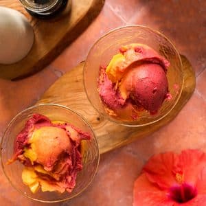 Overhead view of thick and creamy Ninja Creami tropical sorbet with vibrant mango and dragon fruit swirls scooped into two glasses, served beside a red hibiscus flower.