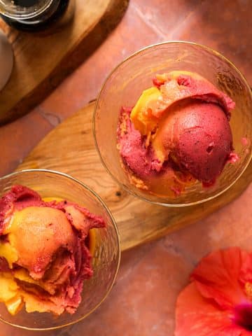 Overhead view of thick and creamy Ninja Creami tropical sorbet with vibrant mango and dragon fruit swirls scooped into two glasses, served beside a red hibiscus flower.